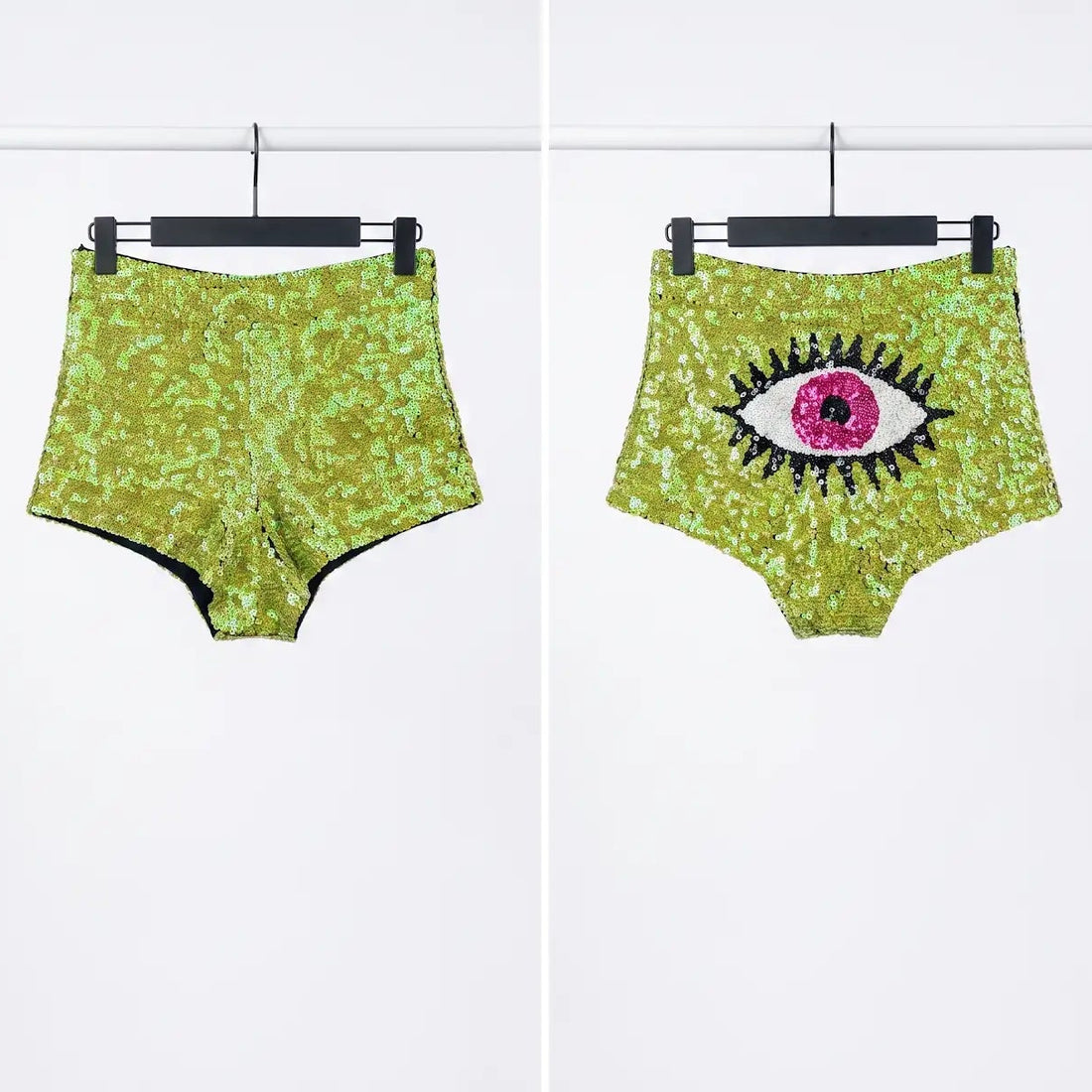 Two pairs of high-waisted, sparkly magenta-purple sequin shorts displayed side by side on black hangers against a light gray background. The left pair has a solid sequin texture, and the right pair features a large black and white eye graphic on the front.